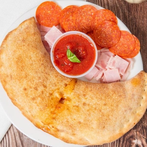 Golden-brown calzone with sides of pepperoni, diced ham, and a cup of marinara sauce.