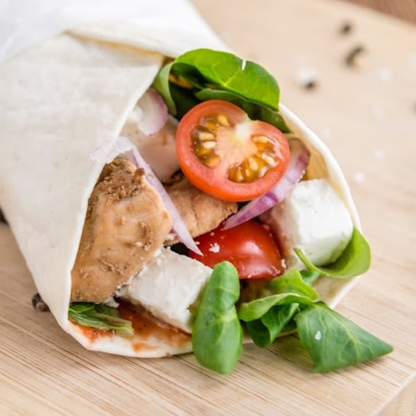 A chicken wrap with tomatoes, red onion, feta cheese, and spinach on a wooden board.