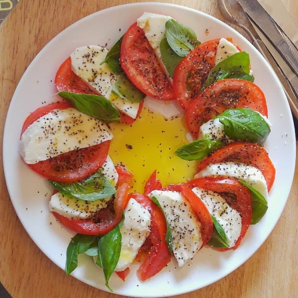 Caprese salad with sliced tomatoes, mozzarella, basil, olive oil, and black pepper on a white plate.